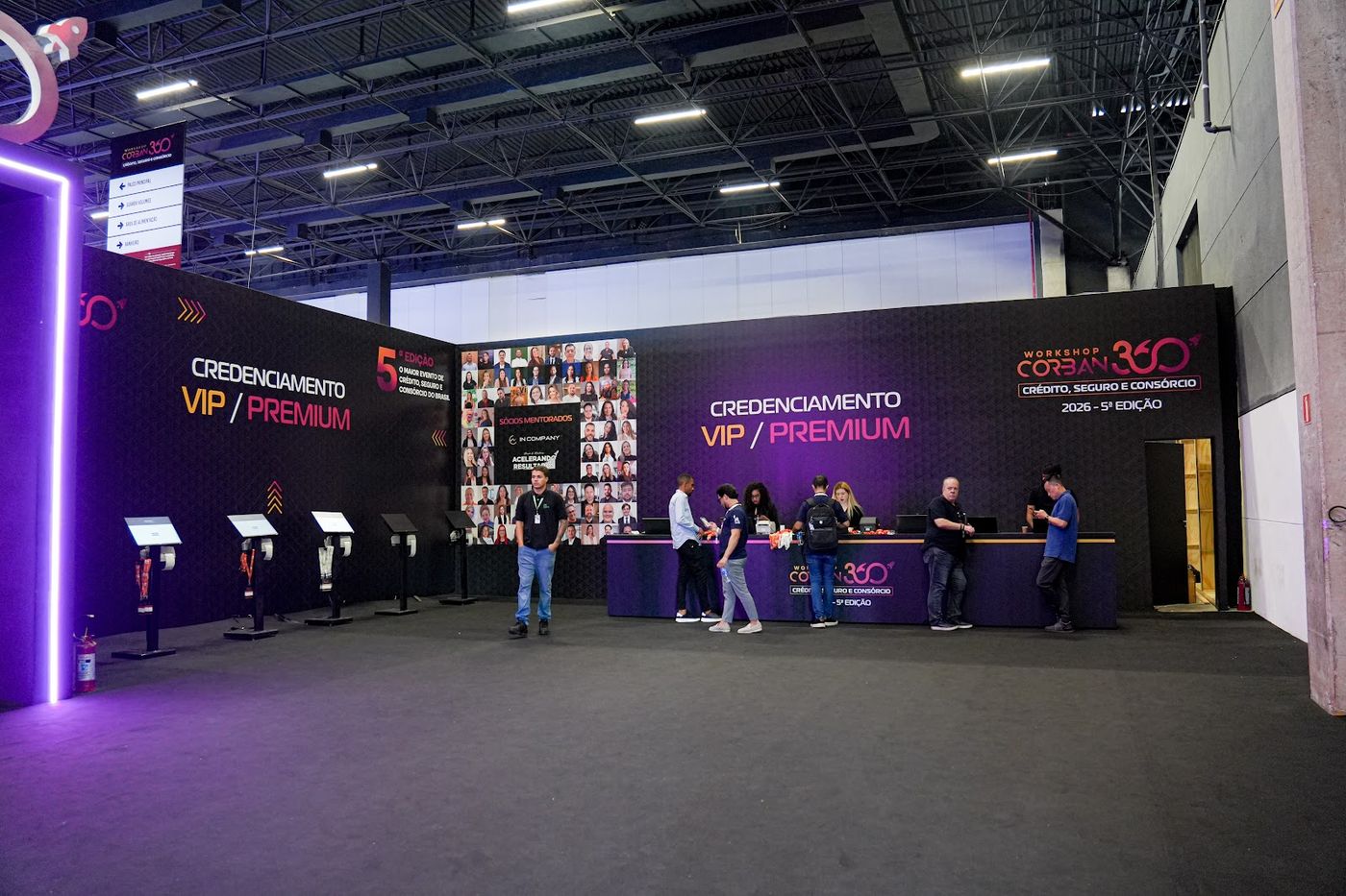 Balcão de credenciamento com painel cenográfico iluminado e totens digitais em evento corporativo