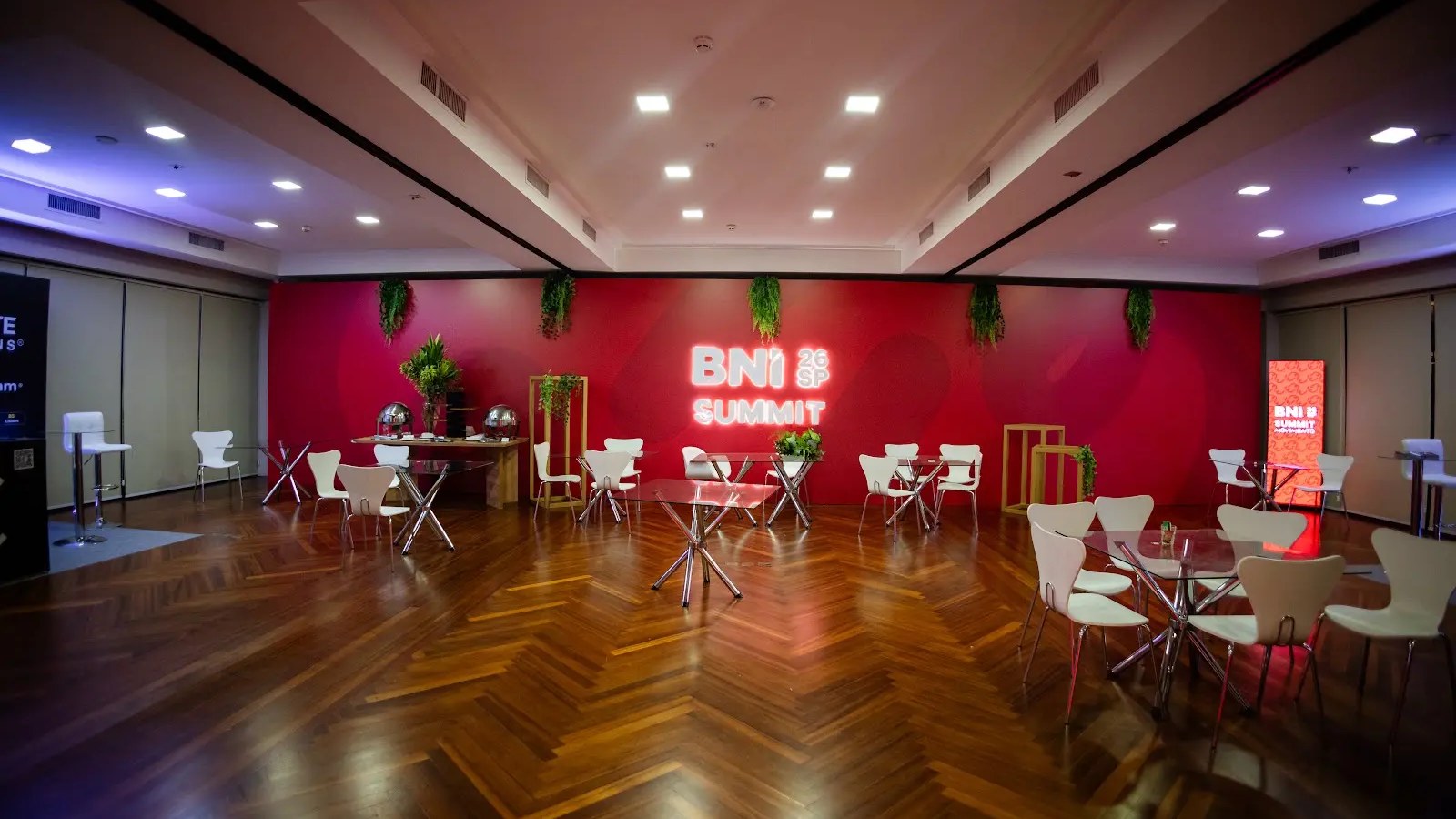 Espaço de networking do BNI Summit 2026 com piso em chevron, backdrop vermelho com letreiro neon e mesas elegantes brancas