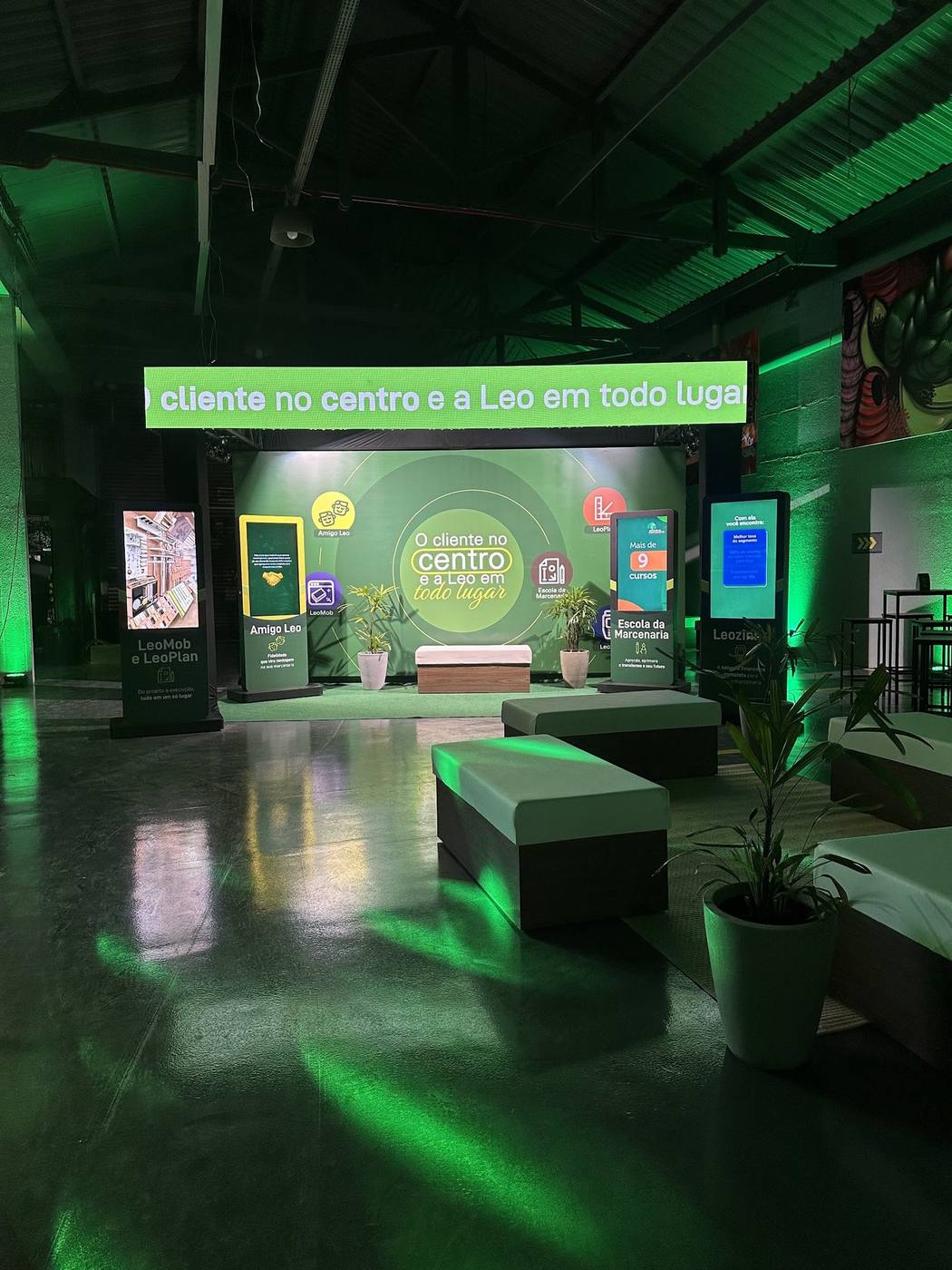 Stand com painéis digitais, totens interativos e lounge iluminado em verde na ativação Léo Madeiras