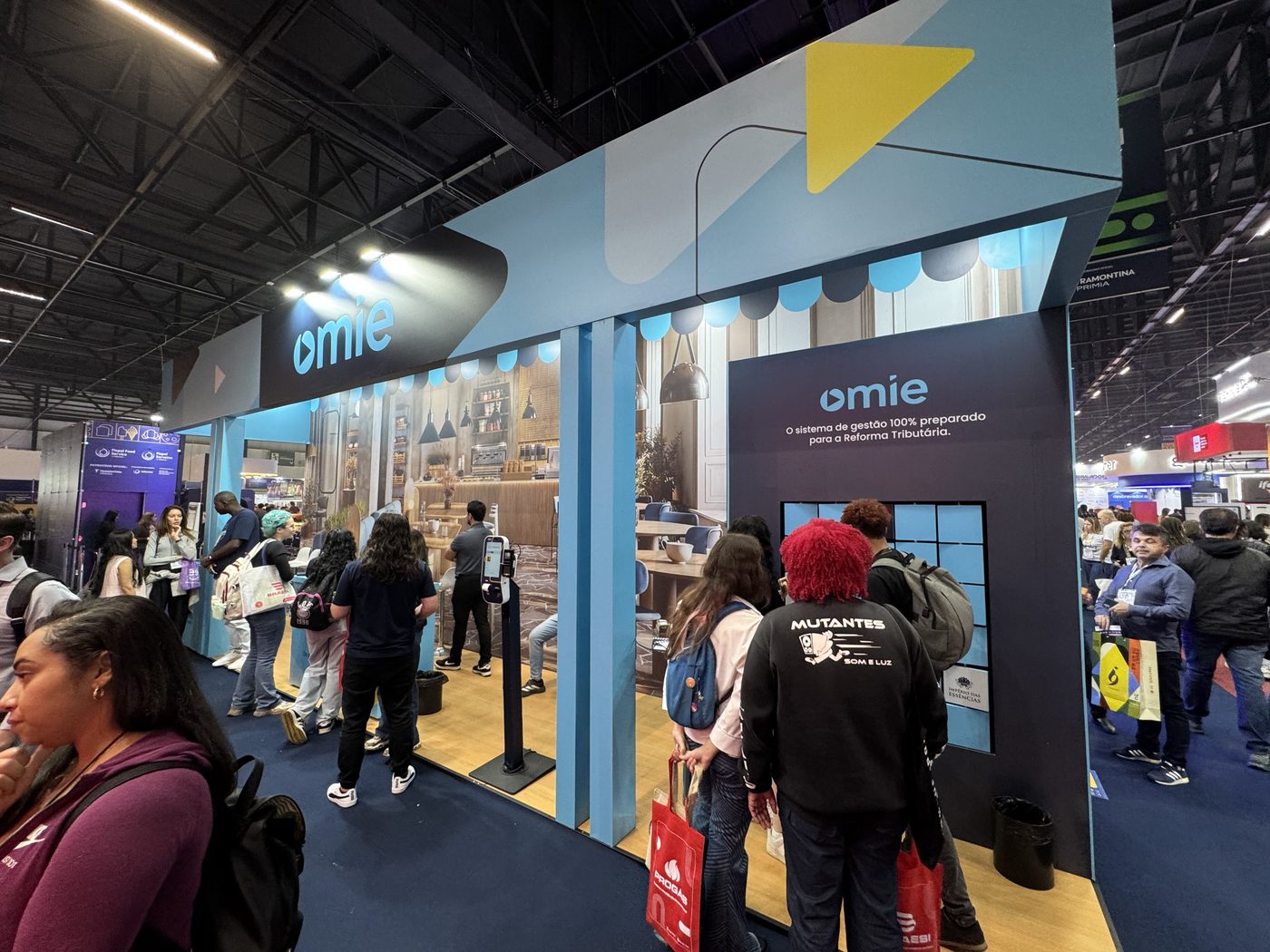 Estande da Omie na Fispal Food Service com testeira azul-marinho e turquesa e cenário de cafeteria-padaria ao fundo